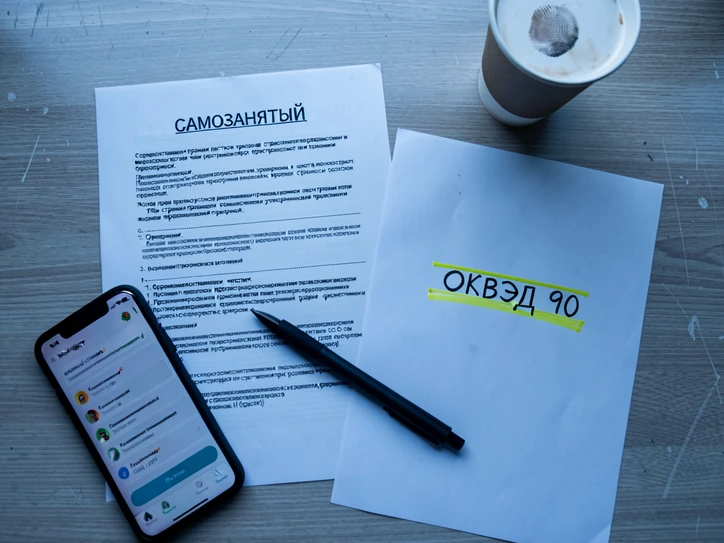 A flat lay top-down shot of a desk with a Russian self-employed registration printout labeled "САМОЗАНЯТЫЙ" on the left, a separate sheet with a highlighted line reading "ОКВЭД 90" in yellow marker on the right, a smartphone showing the «Мой налог» app interface in the lower left corner, a black pen resting diagonally across the papers, and a paper coffee cup with a fingerprint smudge near the top edge. Soft cool daylight from a window above the frame, even and shadowless, gives a clean documentary feel; the wood-grain desk underneath is matte and slightly worn. Sober, administrative, editorial photo atmosphere.