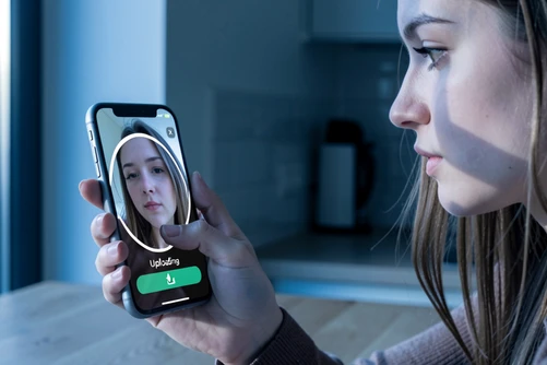 A young woman holds a smartphone showing her own selfie as her thumb presses an upload button inside a face swap app. She sits at a kitchen table beside a tall window. The phone screen displays a circular progress ring and the white sans-serif word "Uploading". Soft cool morning light enters from the left and falls across her cheek and the glossy phone surface. The atmosphere is calm yet quietly uncertain.