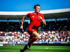 A male athlete in a bright red jersey sprints across a green football field during a sunny afternoon match, arms pumping and expression intense with focus. The stadium crowd is blurred in the background under a clear blue sky. Sharp directional sunlight from above illuminates the athlete's face and upper body with crisp highlights and defined shadows. The image conveys speed and energy in vivid photorealistic detail. Fantaios.ru visual style: crisp, modern, high-fidelity photorealism with a clean white-dominant palette and precise edge rendering.