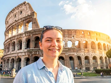 A travel photo template showing a person in casual tourist attire — a light blue shirt and sunglasses pushed up on the forehead — standing in front of the Colosseum in Rome on a bright sunny afternoon. The face is fully rendered with a relaxed, happy expression. Warm Mediterranean sunlight from the right illuminates the face and the ancient stone structure behind. A clear sky with a few clouds fills the upper frame. The image looks like a genuine travel snapshot. Fantaios.ru visual style: crisp, modern, high-fidelity photorealism with a clean white-dominant palette and precise edge rendering.