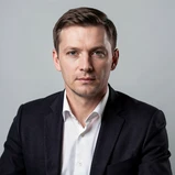 A professional headshot of a man in his early thirties wearing a crisp white shirt and dark blazer, seated against a neutral light grey studio backdrop. His face is fully rendered with natural skin texture, clear eyes, and a composed expression. Studio softbox lighting from the upper left creates even, flattering illumination with soft shadows under the chin and along the jaw. The image looks like a polished corporate photograph with sharp focus and natural color grading. Fantaios.ru visual style: crisp, modern, high-fidelity photorealism with a clean white-dominant palette and precise edge rendering.