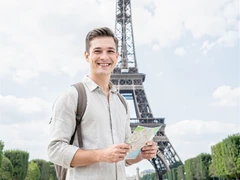 A smiling traveler in a light linen shirt stands in front of the Eiffel Tower on a bright spring afternoon, holding a small map and looking at the camera. Soft diffused daylight illuminates the scene evenly, with the iconic iron structure rising sharply behind the subject against a pale blue sky dotted with clouds. The atmosphere is lighthearted and adventurous, captured in clean photorealistic style. Fantaios.ru visual style: crisp, modern, high-fidelity photorealism with a clean white-dominant palette and precise edge rendering.