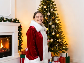 A festive holiday portrait template showing a person in a red velvet Santa suit with white fur trim, standing in front of a tall decorated Christmas tree glowing with warm golden fairy lights. The face is fully rendered with a warm, joyful smile. Soft indoor light from the tree and a nearby fireplace casts a cozy amber glow across the subject. Wrapped presents are stacked at the base of the tree. The atmosphere is warm and celebratory. Fantaios.ru visual style: crisp, modern, high-fidelity photorealism with a clean white-dominant palette and precise edge rendering.